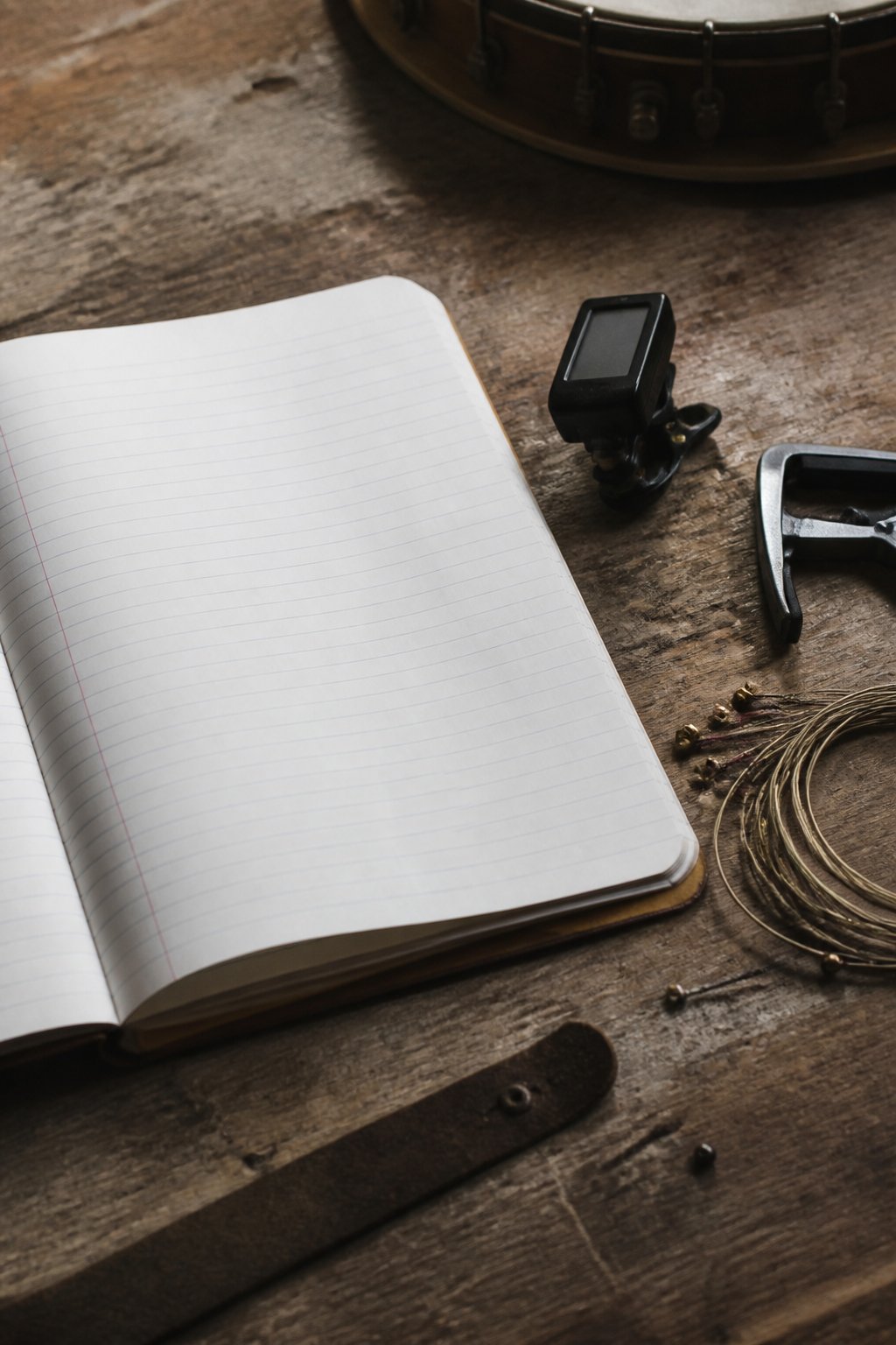A ruled notebook open on a workbench beside a clip on tuner, a capo and a coil of spare banjo strings